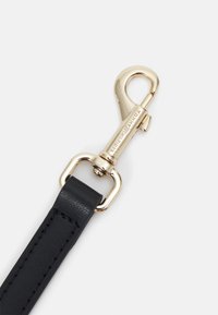 Mousqueton pivotant en métal doré attaché à une sangle en cuir noir avec des coutures visibles sur un fond clair.