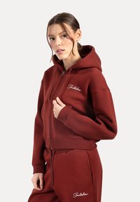 Czerwony hoodie na zamek z przednią kieszenią, ściągaczami i wyszywanym logo. Wykonany z miękkiego materiału w luźnym kroju.