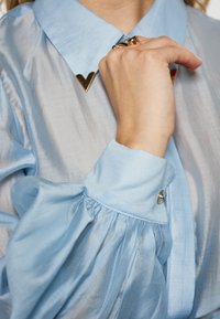 Blusa azzurra chiara con collo strutturato, accentatura geometrica dorata al collo e maniche arricciate con dettaglio a bottone. Texture morbida.