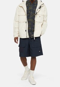 Vit, vattentät jacka med stora framfickor över en mörkt mönstrad skjorta, kombinerad med marinblå cargoshorts och vita sneakers.