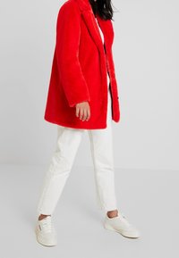 Abrigo rojo de piel sintética con cuello con solapa, cierre frontal de botones y bolsillos laterales, combinado con vaqueros blancos y zapatillas blancas.