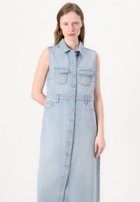 Denim ermeløs kjole i lyseblå med knappesfront, to brystlommer og en tilpasset midje. Myk tekstur, kne-lengde.