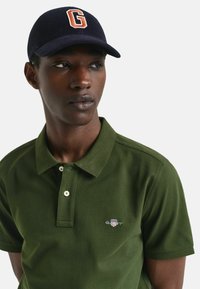 Jongeman met een donkerblauwe pet met de letter G en een olijfgroene polo met een klein logo op de borst, die opzij kijkt.