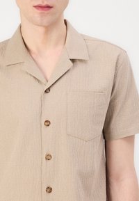 Camisa de botón de manga corta en color beige con una superficie texturizada, un bolsillo en el pecho y cinco botones marrones. Presenta un diseño de cuello clásico.