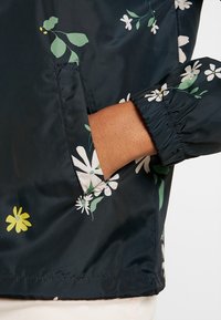Kurtka w kwiatowy wzór w ciemnym granacie z zielonymi liśćmi i białymi stokrotkami. Posiada elastyczne mankiety oraz kieszeń z boku. Gładki, lekki materiał.