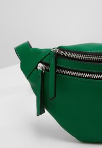Sac banane en cuir texturé vert avec deux fermetures éclair argentées et de longues tirettes en cuir sur fond blanc.