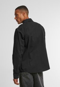 Black button-up shirt with a collar, made of smooth fabric. Features long sleeves and a rounded hem, paired with dark denim jeans.