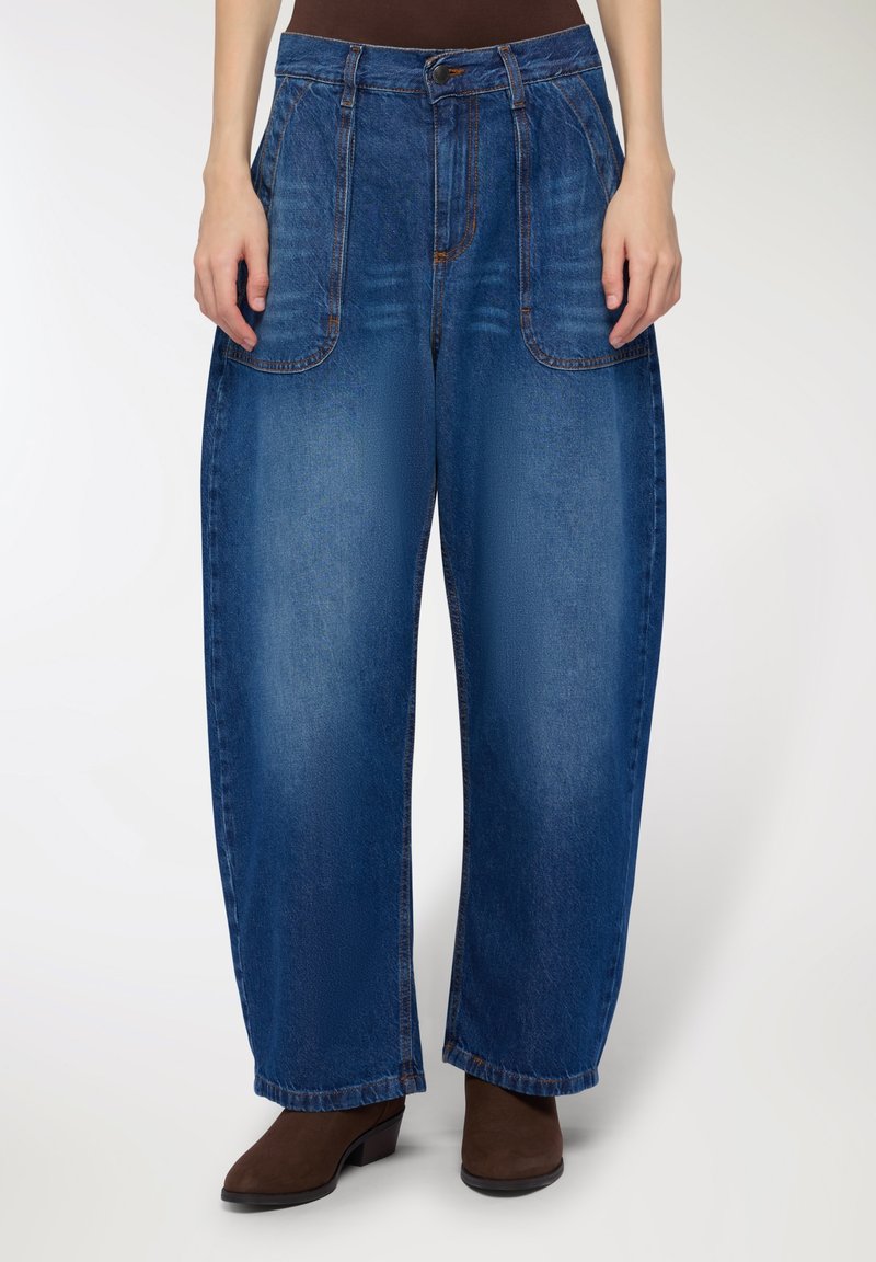 Jeans in denim blu largo con vita alta, tasche frontali e una texture liscia, indossati con stivali alla caviglia marroni con tacco.