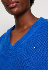 Pull tricoté bleu avec un col en V. Tissu texturé et petite écusson logo en rouge, blanc et bleu sur le côté gauche.