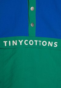 Chemise à manches courtes avec des panneaux bleu et vert, boutons-pressions en métal, et logo "TINY COTTONS" en blanc sur la section verte.