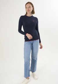 Marineblaues Langarmshirt mit rotem Herz-Logo, kombiniert mit hellblauen geraden Jeans und weißen Sneakers. Einfaches, lässiges Design, aus Baumwollmaterial.