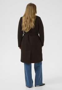 Abrigo marrón texturizado con cinturón, con una abertura en la parte posterior y mangas largas, combinado con unos jeans azul de pierna ancha y botas negras hasta el tobillo.