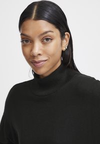 Pulover negru cu guler turceasc ribbed, având un guler strâns și o textură moale. Modelul are părul lung și închis la culoare și poartă cercei mici din aur.