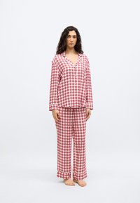 Γυναίκα που στέκεται ξυπόλητη φορώντας κόκκινο και λευκό καρό πουκάμισο με κουμπιά και μακριά μανίκια, μαζί με παντελόνι που ταιριάζει, μπροστά σε λευκό φόντο.