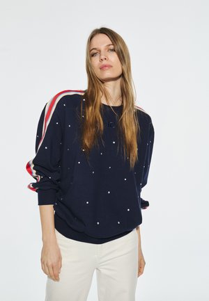 Sudadera azul marino con adornos perlados blancos, detalles en rayas rojas y crema en los hombros, corte relajado, cuello redondo y puños acanalados.