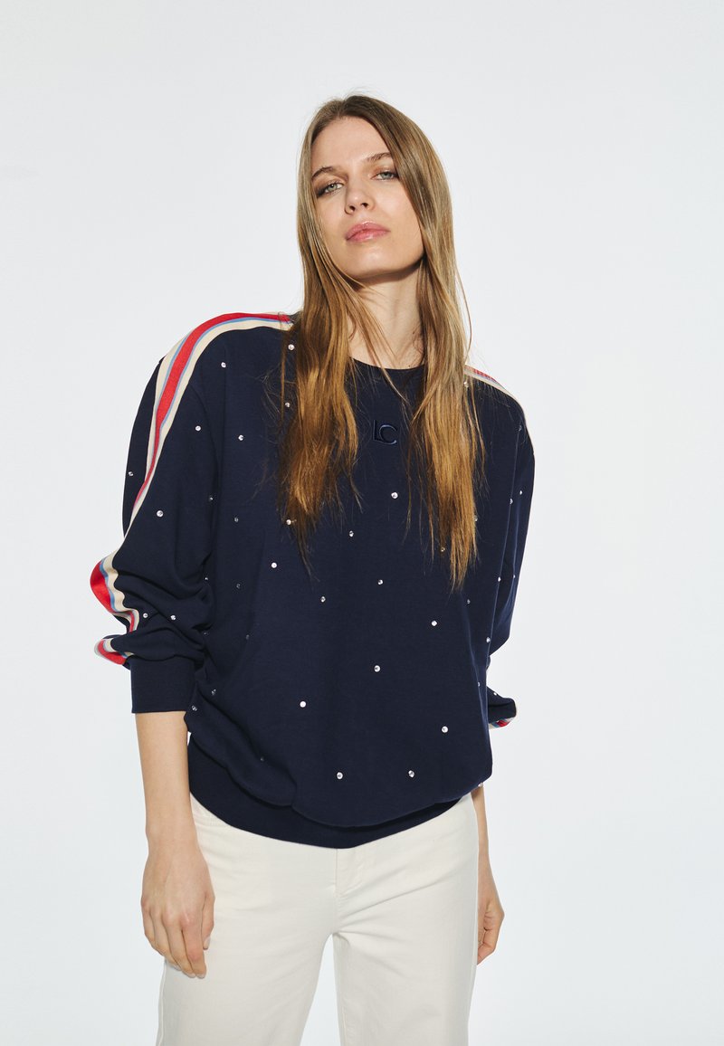 Marinblå sweatshirt med vita pärlliknande dekorationer, randiga röd och gräddfärgade detaljer på axlarna, avslappnad passform, rund hals, ribbad mudd.