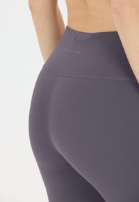 Höga, figurnära leggings i mörklila tyg med en slät textur, som har diskret branding på baksidan.