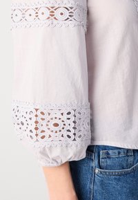 Balta blūze ar uzpūstām piedurknēm, kurai ir izsmalcināta mežģīņu apdare pie aprocēm. Audumam ir maiga, gluda tekstūra un nedaudz caurspīdīga.