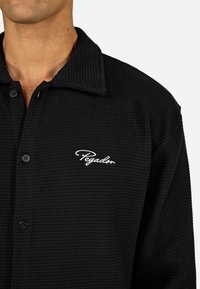 Svart vevd skjorte med krage, med teksturert stoff, knappet front og hvit "Pegador" brodering på brystet.