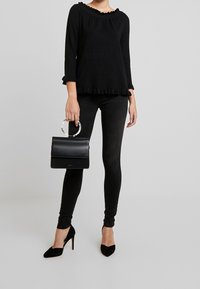 Personne portant un haut noir à volants, un jean skinny noir, des talons hauts pointus noirs, tenant un sac à main noir avec une anse ronde blanche.