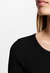Top en maille côtelée noire avec un col rond, présentant une fine texture verticale ; le matériau semble extensible et ajusté.