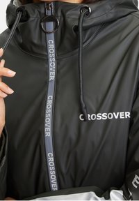 Černá nepremokavá bunda s vysokým límcem. Má šedé logo „CROSSOVER“ a zip, elastický stahovací šňůrku a hladkou texturu.