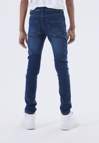 Tievās zilās džinsēs ar vidēju jostas līmeni, nelielu mazgāšanu un divām aizmugurējām kabatām. Valkā ar baltajiem augstajiem sporta apaviem.