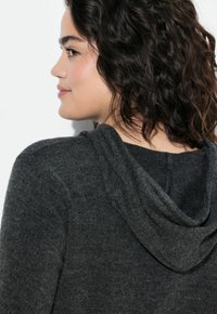 Pull gris à capuche avec un tricot texturé et une capuche ample. Le tissu semble doux et confortable, idéal pour un usage décontracté.