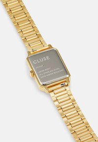 Montre-bracelet en acier inoxydable doré avec un boîtier rectangulaire, affichant un design minimaliste, un bracelet à maillons texturé et un dos gravé.