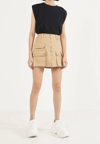 Kaki cargorock med två framfickor och central dragkedja, parat med en svart ärmlös topp. Bärs med vita klumpiga sneakers.