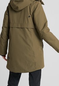 Veste imperméable vert olive avec une finition texturée, capuche et ourlet arrière incliné ; comporte un logo et des fermetures à pression.