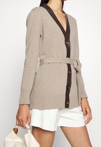 Béžový pletený kardigan s tmavě hnědým výstřihem ve tvaru V, pěti knoflíky vpředu, páskem na zavázání v pase a dlouhými rukávy.