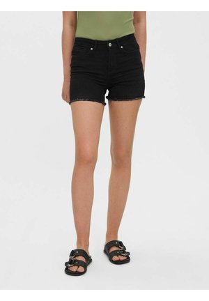 Person iført sorte denimshorts med frynset kant og sorte sandaler med spænder, stående på en ensfarvet lys baggrund.