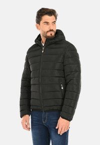 Uomo che indossa un giubbotto a rombi nero con cappuccio e tasche con zip, e jeans blu, mentre guarda di lato su uno sfondo neutro.