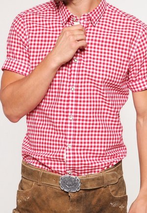 Man past de kraag aan van een rood-wit geruit overhemd met korte mouwen, draagt bruine suède leren broek met decoratieve riemgesp.
