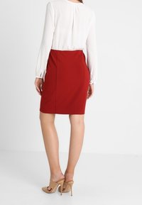 Rode, aansluitende rok met een gladde textuur, gecombineerd met een witte blouse. De outfit wordt aangevuld met beige hoge sandalen.