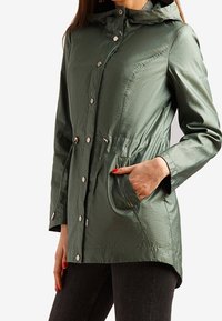Parka w oliwkowym zielonym kolorze, wodoodporna, z kapturem, sznurkiem w talii, guzikami i dwoma przednimi kieszeniami. Miękka, teksturowana tkanina.