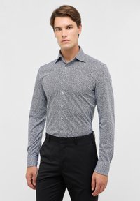 Langarmshirt in een blauw-wit abstract patroon, met een aansluitend ontwerp, standaard kraag en knoopsluiting aan de voorkant. Gecombineerd met zwarte broek.