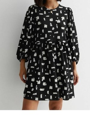 Woman wearing a black dress with white abstract square patterns, long puffed sleeves, and a gathered waist, standing against a plain background.