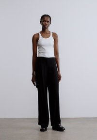 Fekete, bőszárú nadrág fehér trikóval párosítva, egyszerű dizájnt bemutatva. A modell semleges tónusú, minimalista környezetben áll.