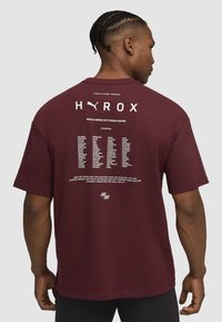 Bordeauxrotes Kurzarmhemd mit großem weißen "HYROX"-Logo auf dem Rücken, darunter die Veranstaltungsdetails und die Namen der Übungen angeordnet.