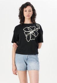 T-shirt noir à manches courtes avec un motif de fleur brodé blanc sur le côté gauche. Le modèle l'associe à un short en denim.