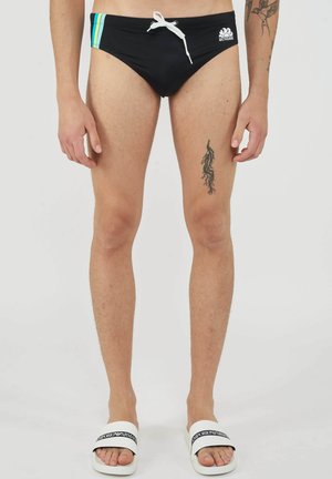 Man draagt zwarte zwembroek met zijstrepen, witte trekkoord, zichtbare dijtattoos en witte Emporio Armani slippers op een effen achtergrond.