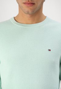 Pull en tricot vert menthe clair avec un col côtelé, présentant un petit patch logo en marine, rouge et blanc sur la zone de la poitrine gauche.