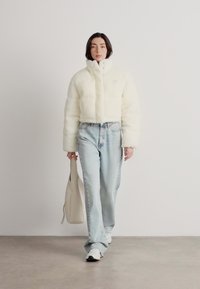 Biela skrátená kabátik s náplňou a vysokým golierom, nosený cez svetlo modré, mierne vyblednuté džínsy. Model má béžovú nákupnú tašku.