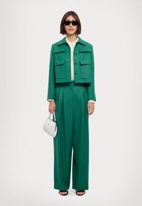 Giacca cropped verde smeraldo con tasche frontali, abbinata a pantaloni larghi. Il modello porta una piccola borsa bianca con un accento argento.