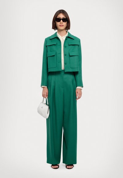 Smaragdgroene cropped jas met voorzakken, gecombineerd met losse broek. Model draagt een kleine witte handtas met een zilveren accent.
