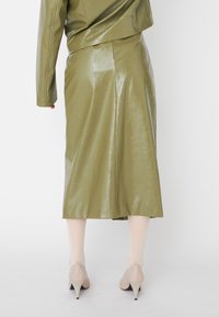 Jupe midi en cuir verni vert olive avec une forme évasée et une fente au dos. Portée avec des talons aiguilles de couleur claire.