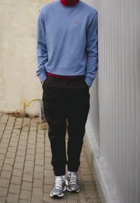 Felpa azzurra con logo rosso, indossata sopra un dolcevita rosso; pantaloni neri a gamba stretta; sneakers grigie con dettagli bianchi e rossi.