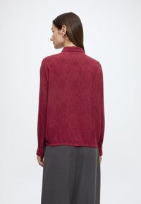 Bordeauxrode blouse met lange mouwen, een getextureerd verticaal gestreept patroon en een kraag. Voorzien van knoopmanchetten en een licht losse pasvorm.